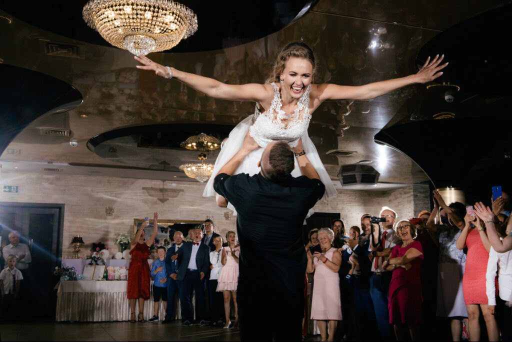 Uśmiechnięta panna młoda w białej sukni, podnoszona przez mężczyznę na tle wiwatującego tłumu gości weselnych w eleganckiej sali z żyrandolami.