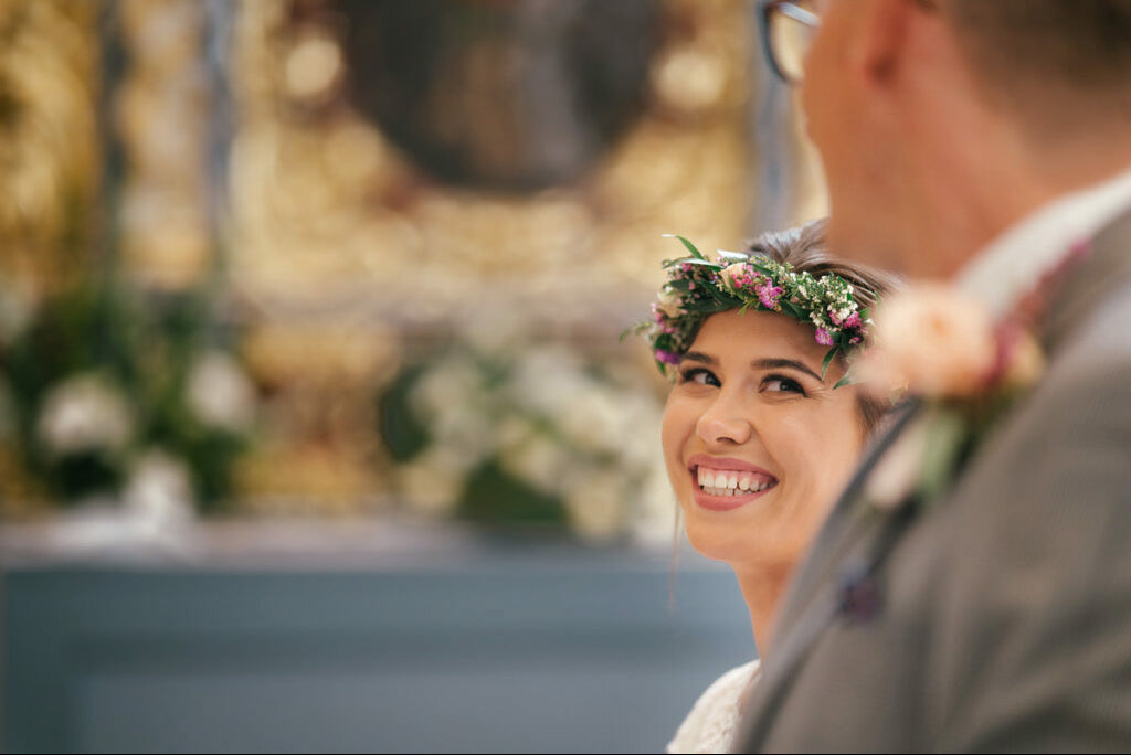 Uśmiechnięta panna młoda w wianku kwiatów, spoglądająca w bok podczas ceremonii ślubnej. W tle rozmyte dekoracje kościelne i fragment sylwetki pana młodego.