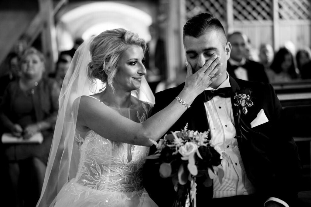 Czarno-białe zdjęcie wzruszonej panny młodej ocierającej łzy panu młodemu podczas ceremonii ślubnej. W tle goście i sklepienie kościoła.