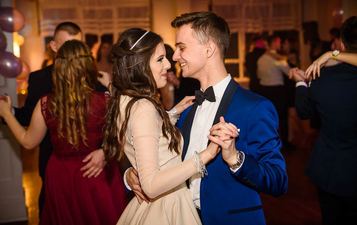 Uśmiechnięta para w tańcu na balu, on w niebieskim smokingu, ona w beżowej sukience, w tle inni tańczący, ciepłe światło, widoczne balony.