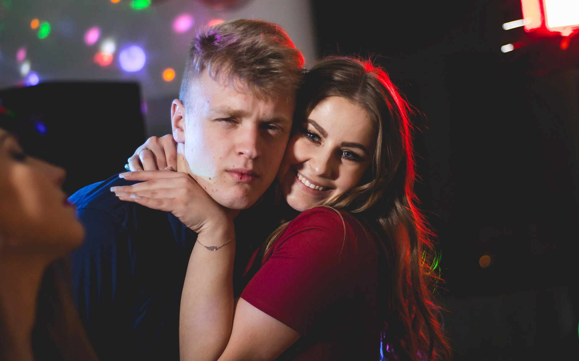 Uśmiechnięta para obejmuje się na tle rozmytych świateł dyskoteki. Kobieta w czerwonej sukience, mężczyzna w ciemnej koszuli. Widoczny fragment twarzy innej osoby.