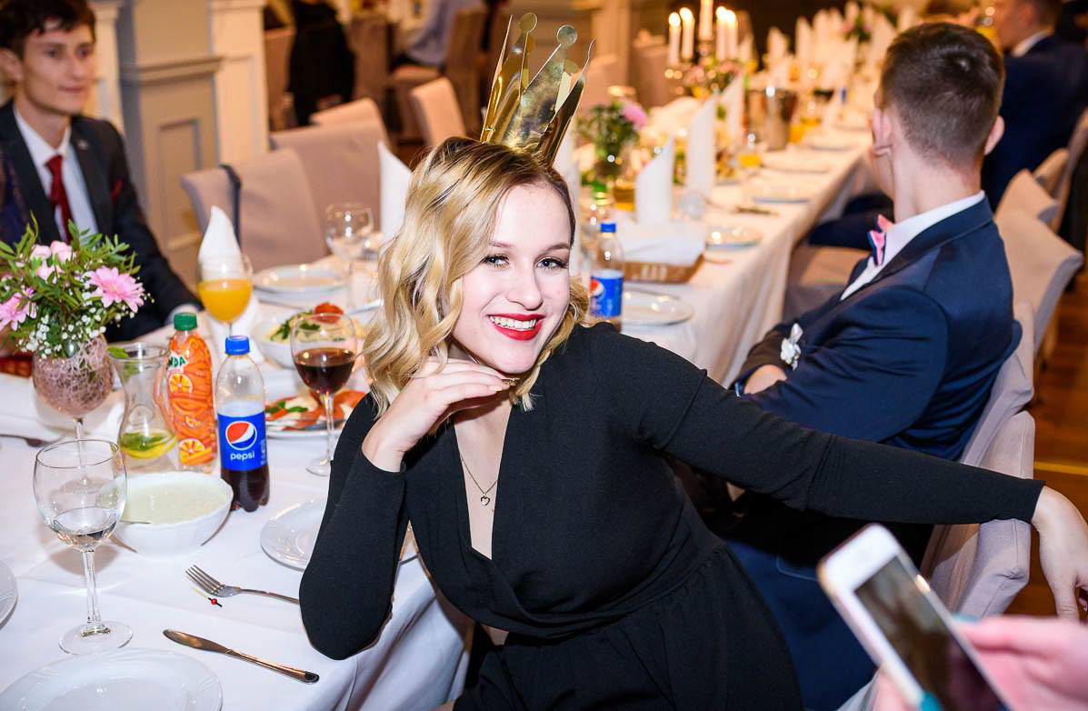 Uśmiechnięta blondynka w koronie siedzi przy stole podczas eventu. Widoczne napoje, jedzenie, dekoracje i inni goście. Zdjęcie z imprezy okolicznościowej.