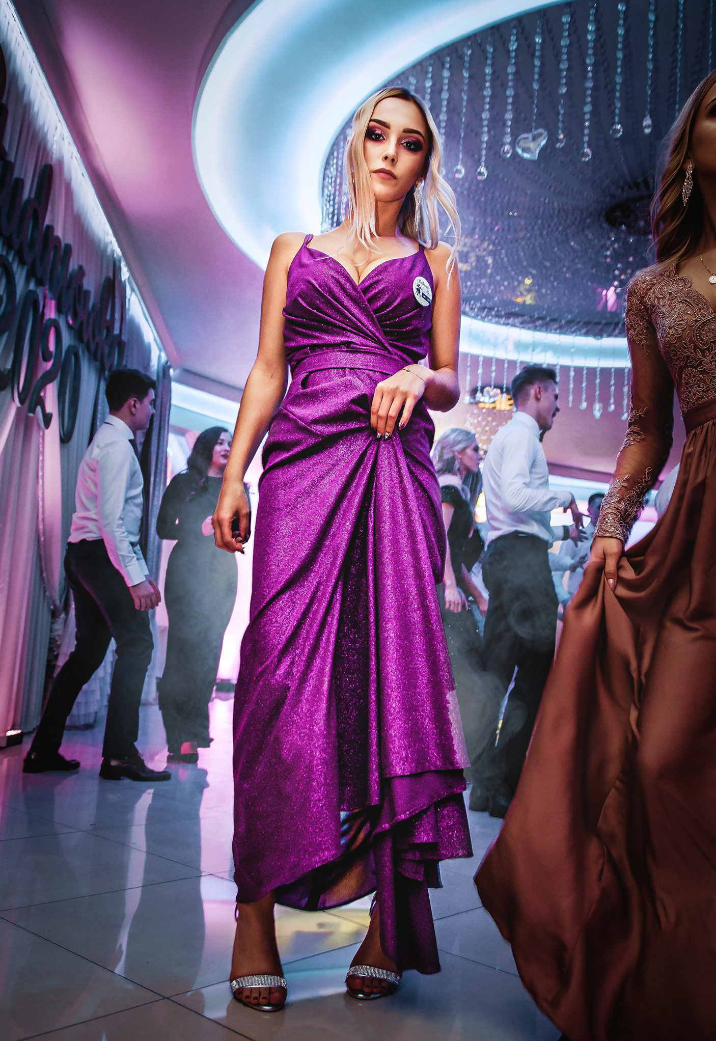 Kobieta w fioletowej sukni na tle sali balowej z dekoracjami i tańczącymi ludźmi. Uwagę przykuwa błyszcząca tkanina sukni i oświetlenie sceniczne.