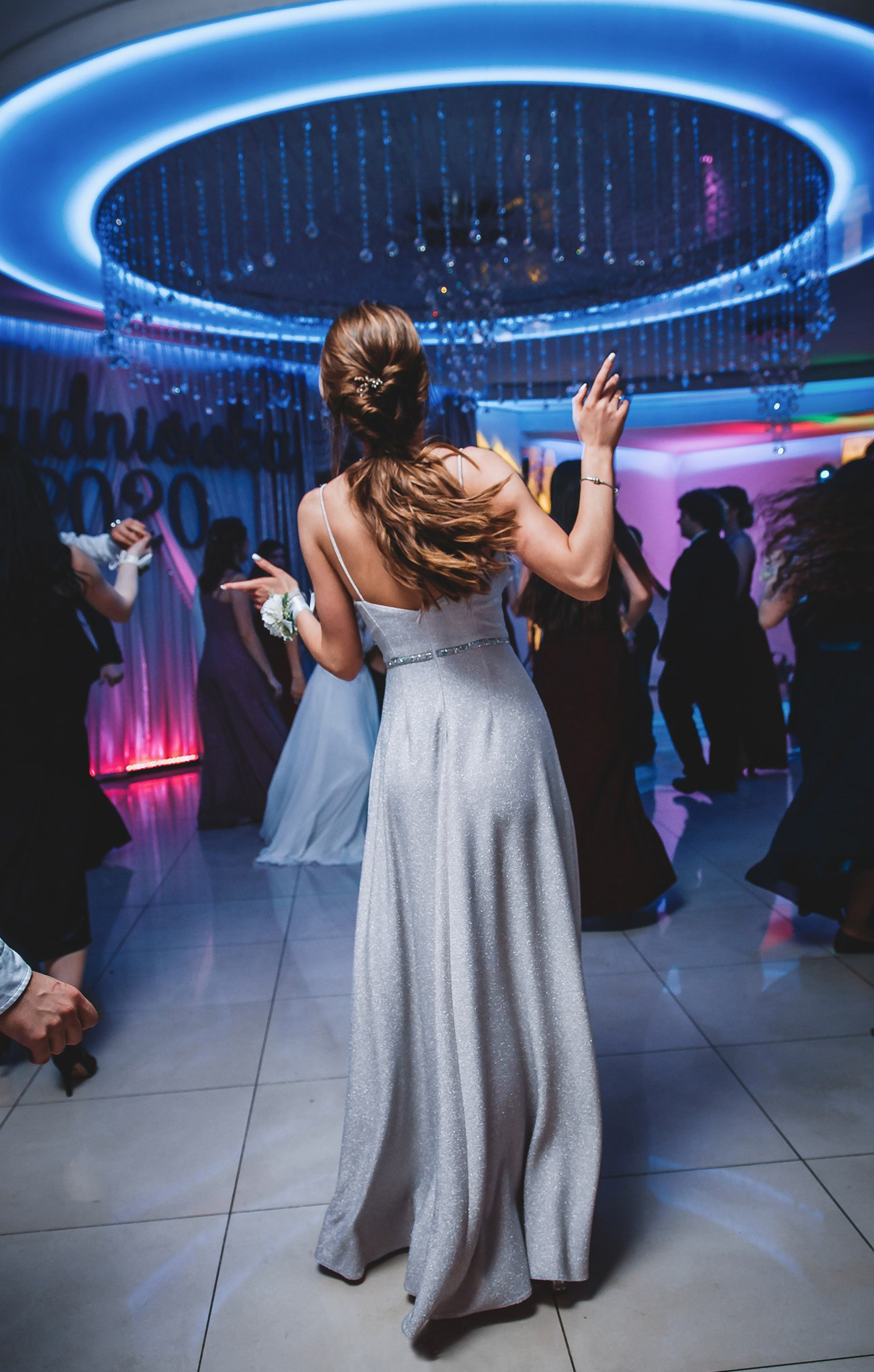 Tancerka w srebrnej sukni na tle rozświetlonej sali balowej z dekoracjami i innymi osobami. Widok z tyłu, dynamiczne ujęcie.