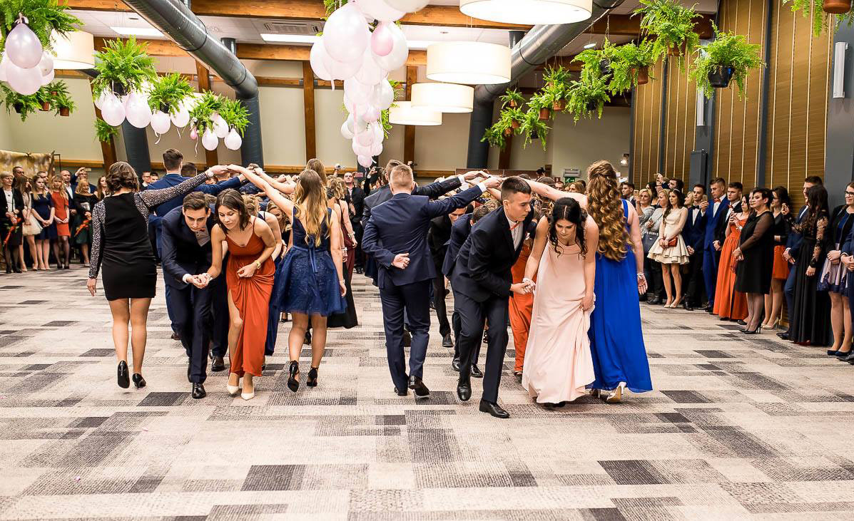 Polonez na studniówce: Młodzież w eleganckich strojach tańczy w rzędzie, tworząc tunel z rąk. Dekoracje z balonów i roślin zwisają z sufitu. Parkiet w geometryczny wzór.