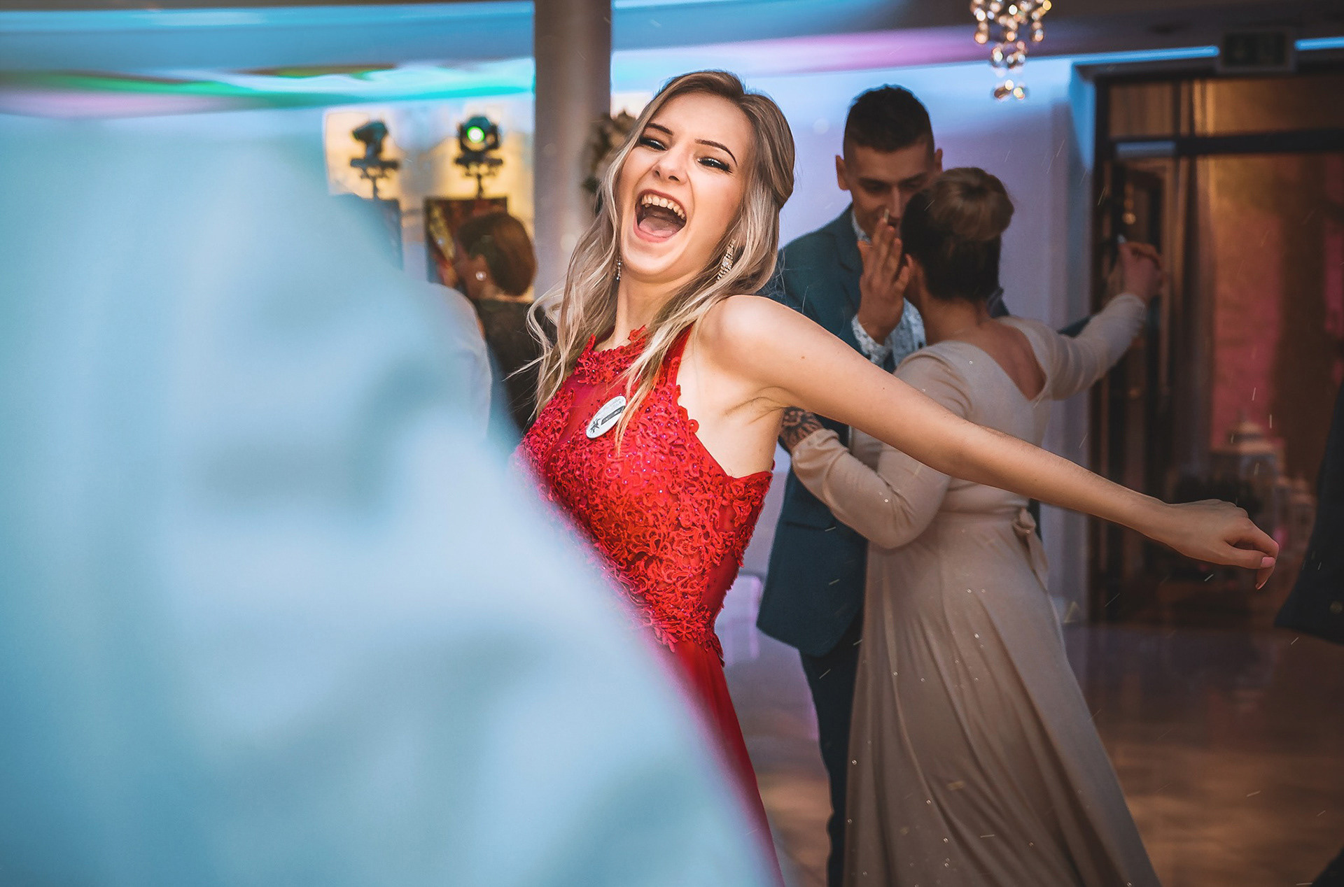 Rozbawiona blondynka w czerwonej sukni tańczy na parkiecie podczas eventu. W tle para tańcząca i oświetlenie sceniczne. Uśmiech, radość i dynamika uchwycone w kadrze.
