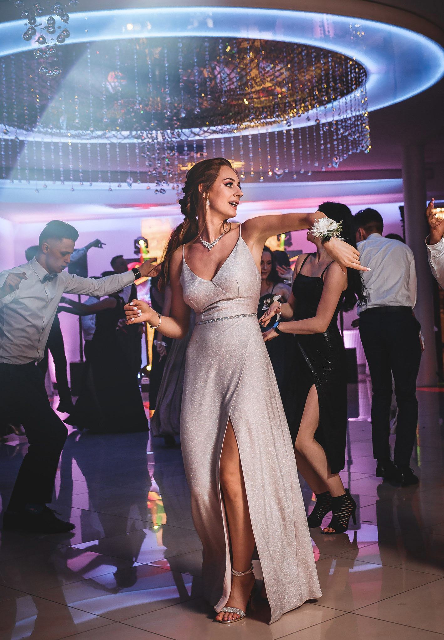 Roztańczona kobieta w srebrnej sukni na parkiecie, otoczona innymi osobami. W tle dekoracyjny żyrandol i efekty świetlne tworzą imprezową atmosferę.