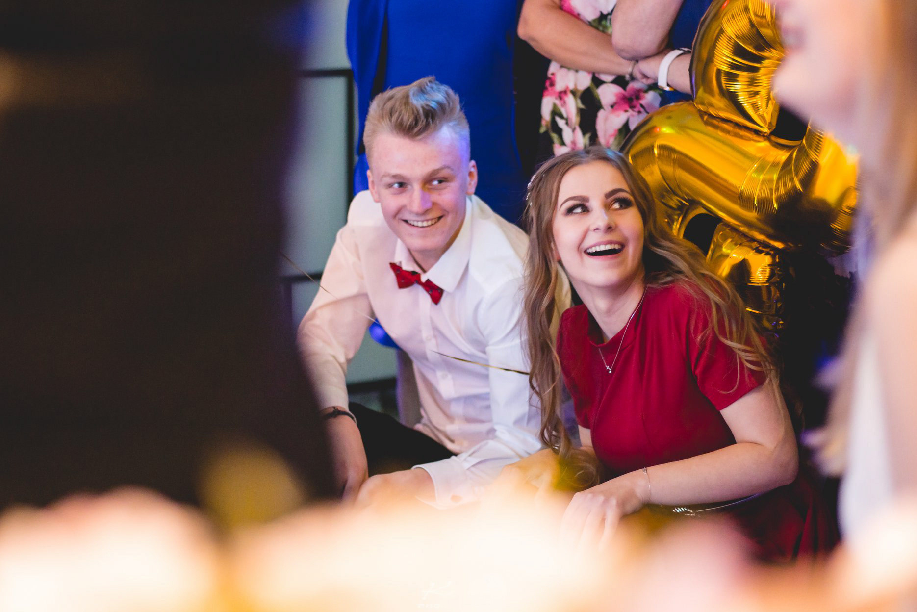Uśmiechnięta para na tle złotych balonów podczas imprezy. Mężczyzna w białej koszuli i muszce, kobieta w czerwonej sukience. Radosna atmosfera, dynamiczne ujęcie.