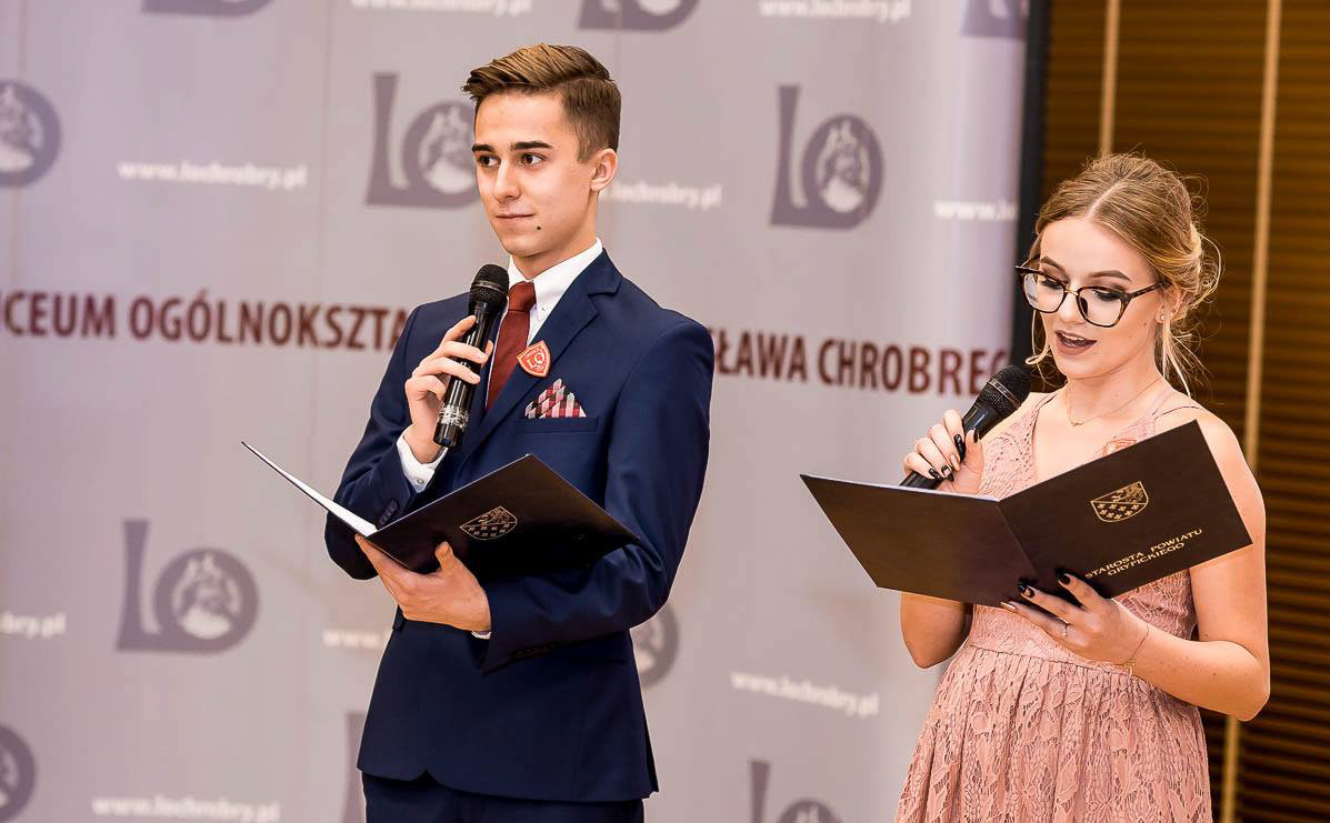 Dwoje młodych ludzi w eleganckich strojach przemawia do mikrofonu podczas uroczystości. W tle widoczne logo i napisy.