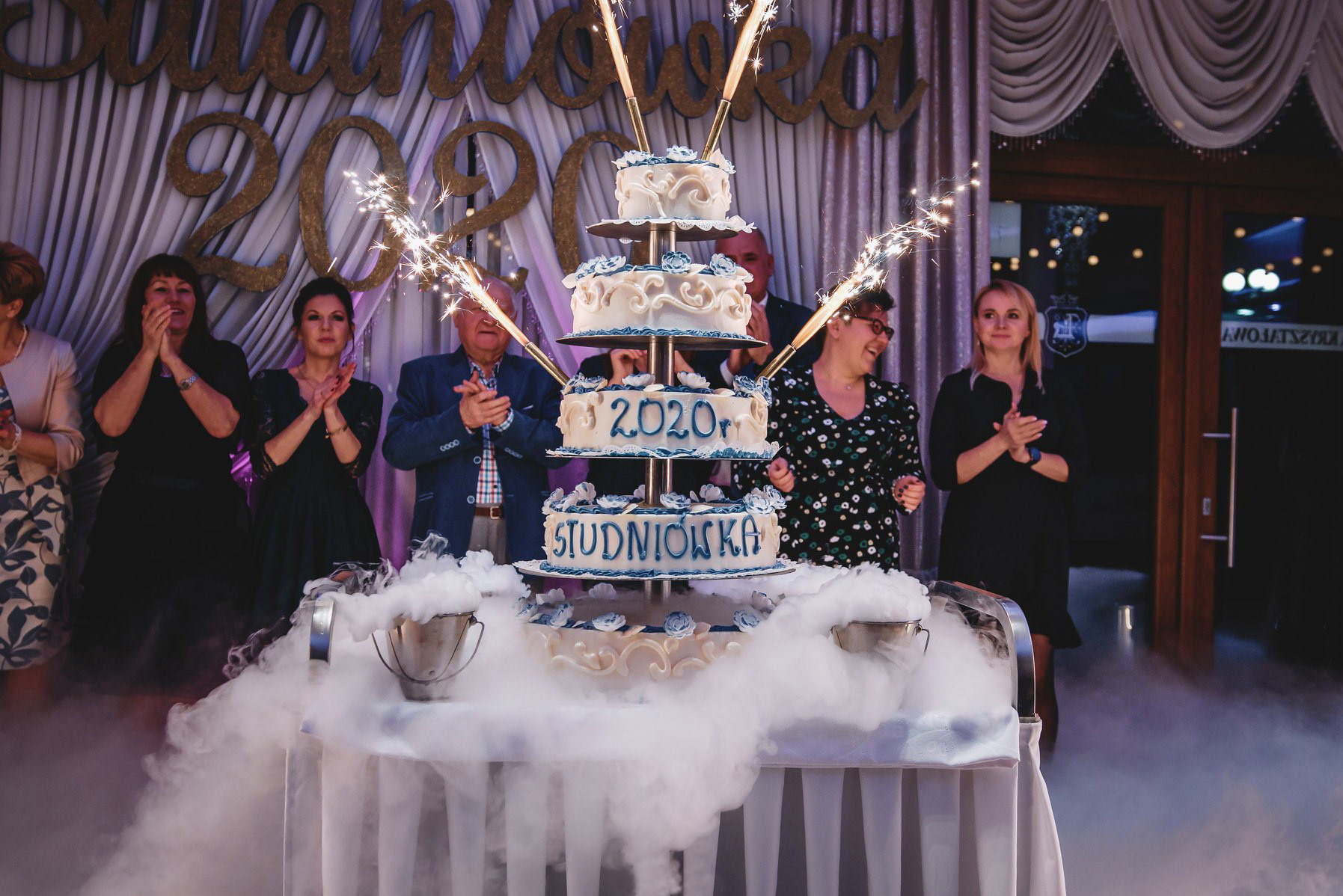 Uroczysty tort studniówkowy z fajerwerkami i efektem dymu, otoczony gośćmi klaszczącymi w dłonie na tle dekoracji z napisem 'Studniówka 2020'.