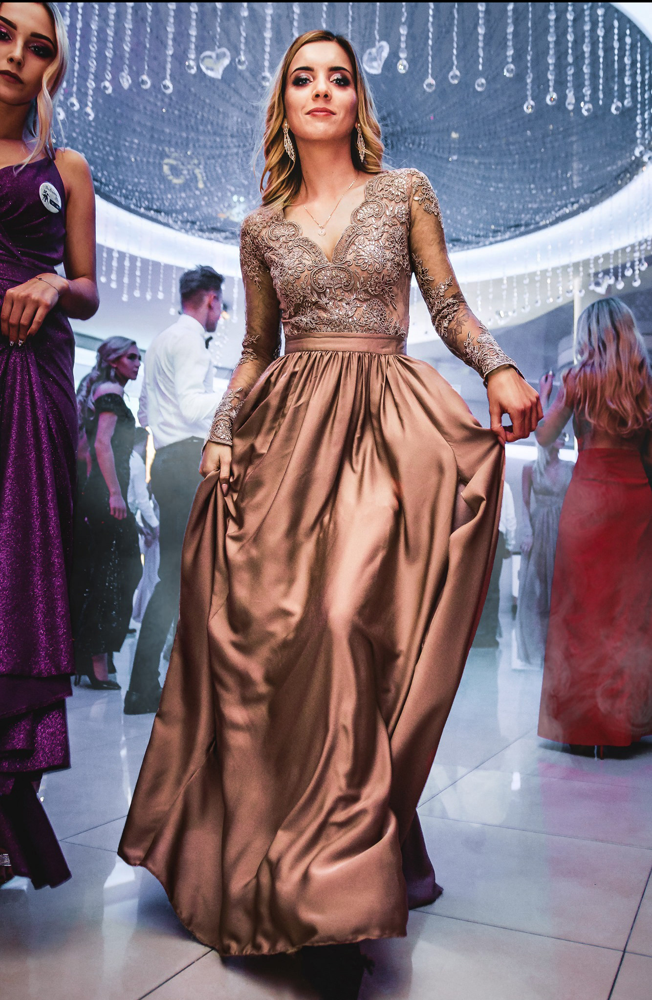 Elegancka kobieta w balowej sukni z koronkową górą, trzymająca materiał sukni, na tle sali balowej z innymi osobami i dekoracyjnym oświetleniem.