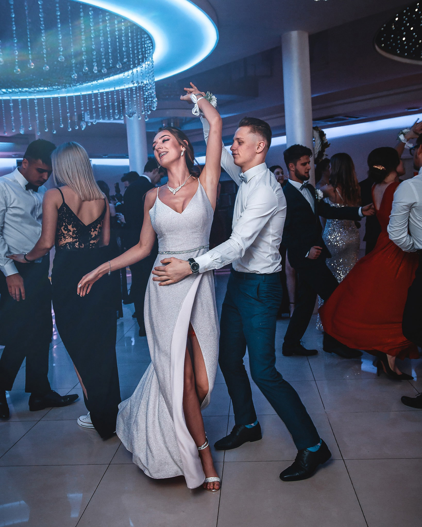 Para tańczy na parkiecie podczas uroczystości. Kobieta w srebrnej sukni z rozcięciem, mężczyzna w białej koszuli i granatowych spodniach. W tle goście i dekoracyjny żyrandol.