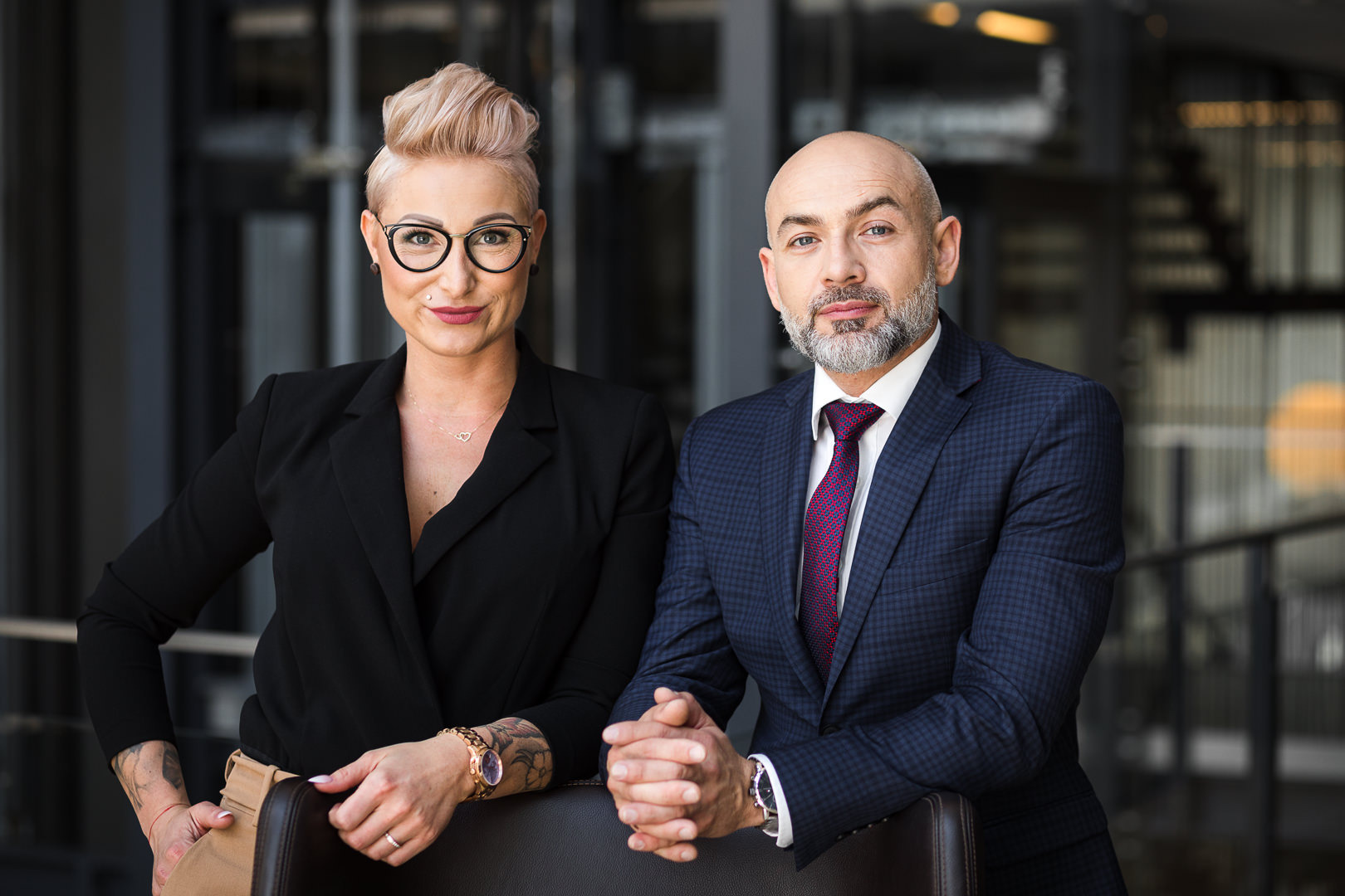 Profesjonalny portret biznesowy: kobieta w czarnej marynarce i mężczyzna w garniturze, oboje z dłońmi splecionymi na oparciu krzesła, na tle nowoczesnego biura.