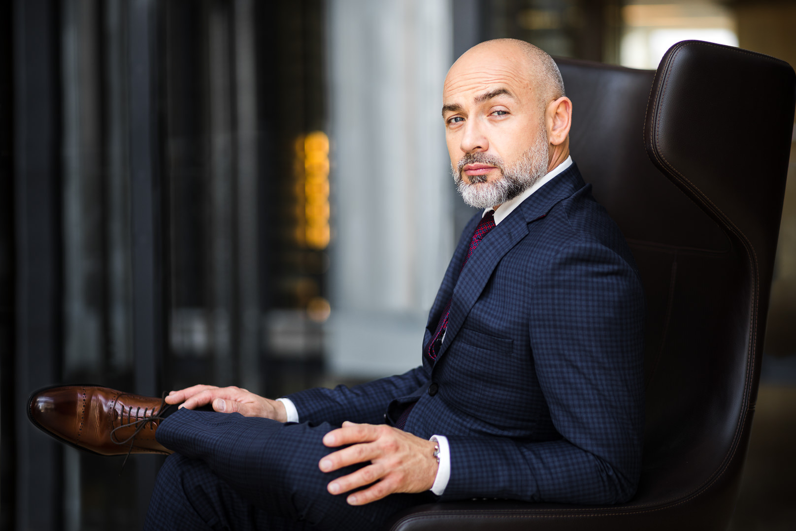 Mężczyzna z brodą w garniturze w kratę siedzi w skórzanym fotelu, spoglądając w obiektyw. Elegancki strój, brązowe buty, stonowane tło. Sesja biznesowa.