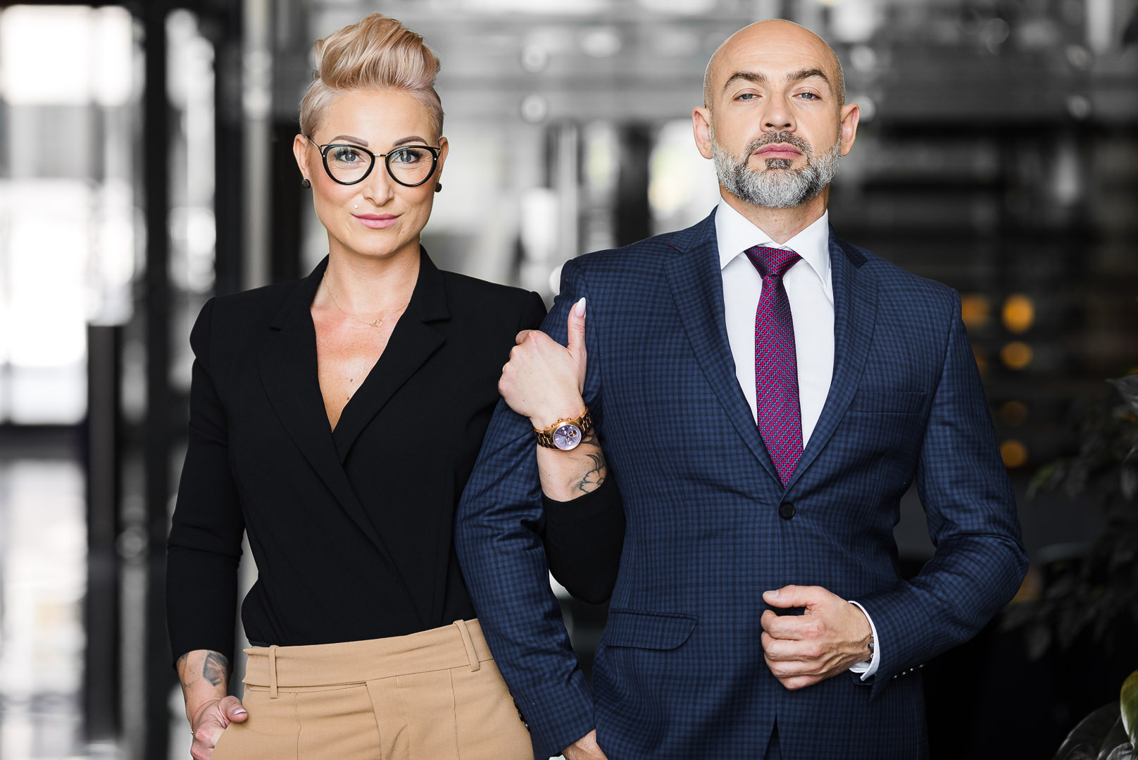Profesjonalny portret biznesowy w Łodzi: elegancka kobieta w okularach opiera rękę na ramieniu mężczyzny w garniturze. Stonowane tło biurowe, oświetlenie studyjne.