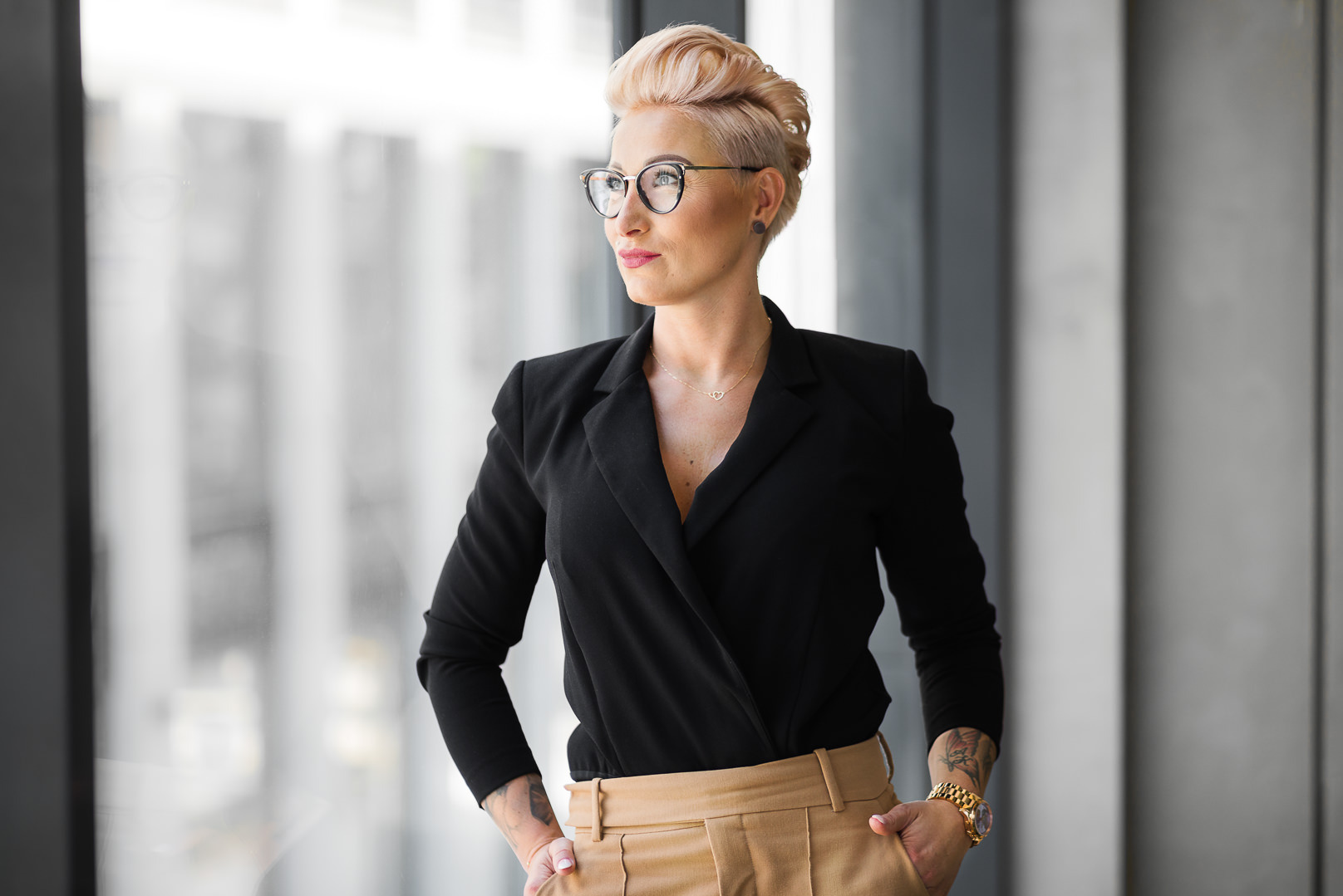 Profesjonalny portret kobiety w okularach, czarnej marynarce i beżowych spodniach, z tatuażami na rękach, stojącej na tle okna w biurze. Krótkie blond włosy.