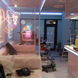 51 Mieczysław Terlecki - Montaż neonów w lokalu 'Candy': neonowe serca i deser za kratą, drabina, narzędzia, osłonięte meble. Prace w toku, specyficzne oświetlenie.