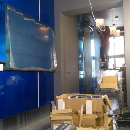 51 Mieczysław Terlecki - Montaż oświetlenia: instalator na drabinie, w tle lampy wiszące, na pierwszym planie niebieska ściana i kartony z materiałami.