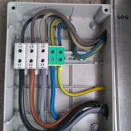 51 Mieczysław Terlecki - Wnętrze skrzynki elektrycznej z podłączonymi przewodami o różnych kolorach i złączkami, zamontowanej na ścianie. Widoczna stara puszka instalacyjna.