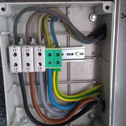 51 Mieczysław Terlecki - Wnętrze puszki elektrycznej z podłączonymi przewodami o różnych kolorach. Widoczne złącza, listwa montażowa i śruby mocujące. Detal instalacji w szarej obudowie.