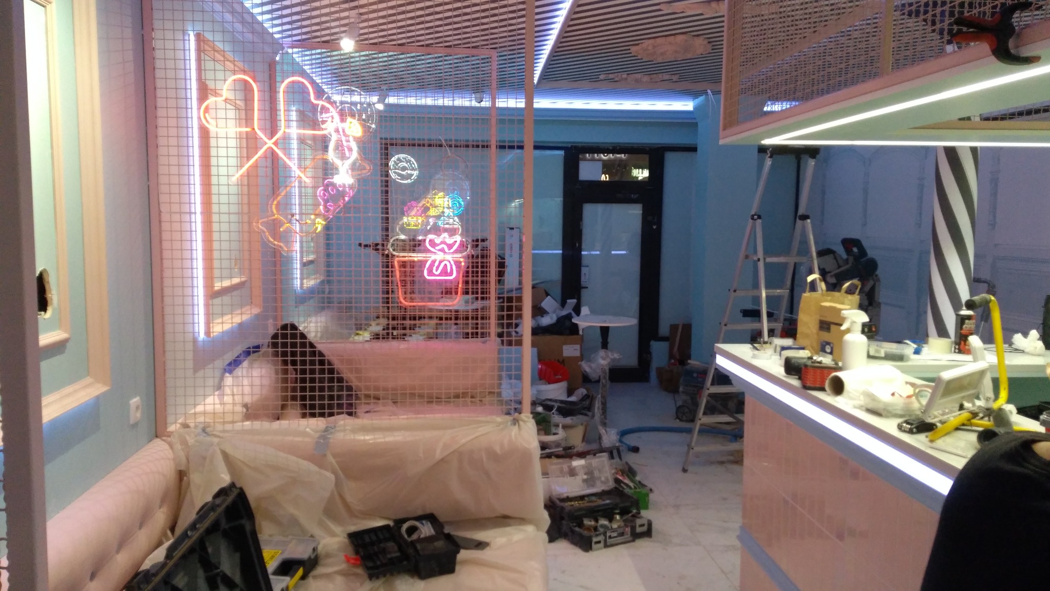 Montaż neonów w lokalu 'Candy': neonowe serca i deser za kratą, drabina, narzędzia, osłonięte meble. Prace w toku, specyficzne oświetlenie.