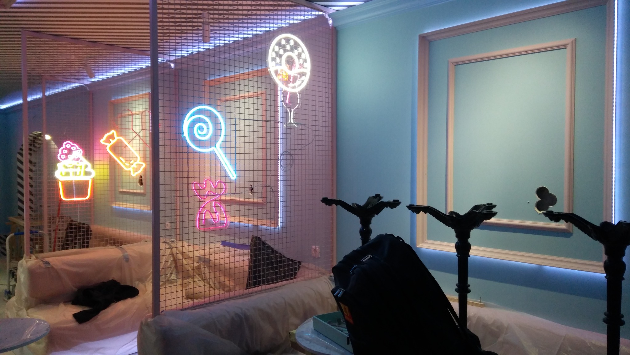 Instalacja neonowa w pastelowych kolorach z motywami słodyczy, widoczna przez metalową siatkę. W tle błękitna ściana z dekoracyjną ramą i oświetleniem LED.