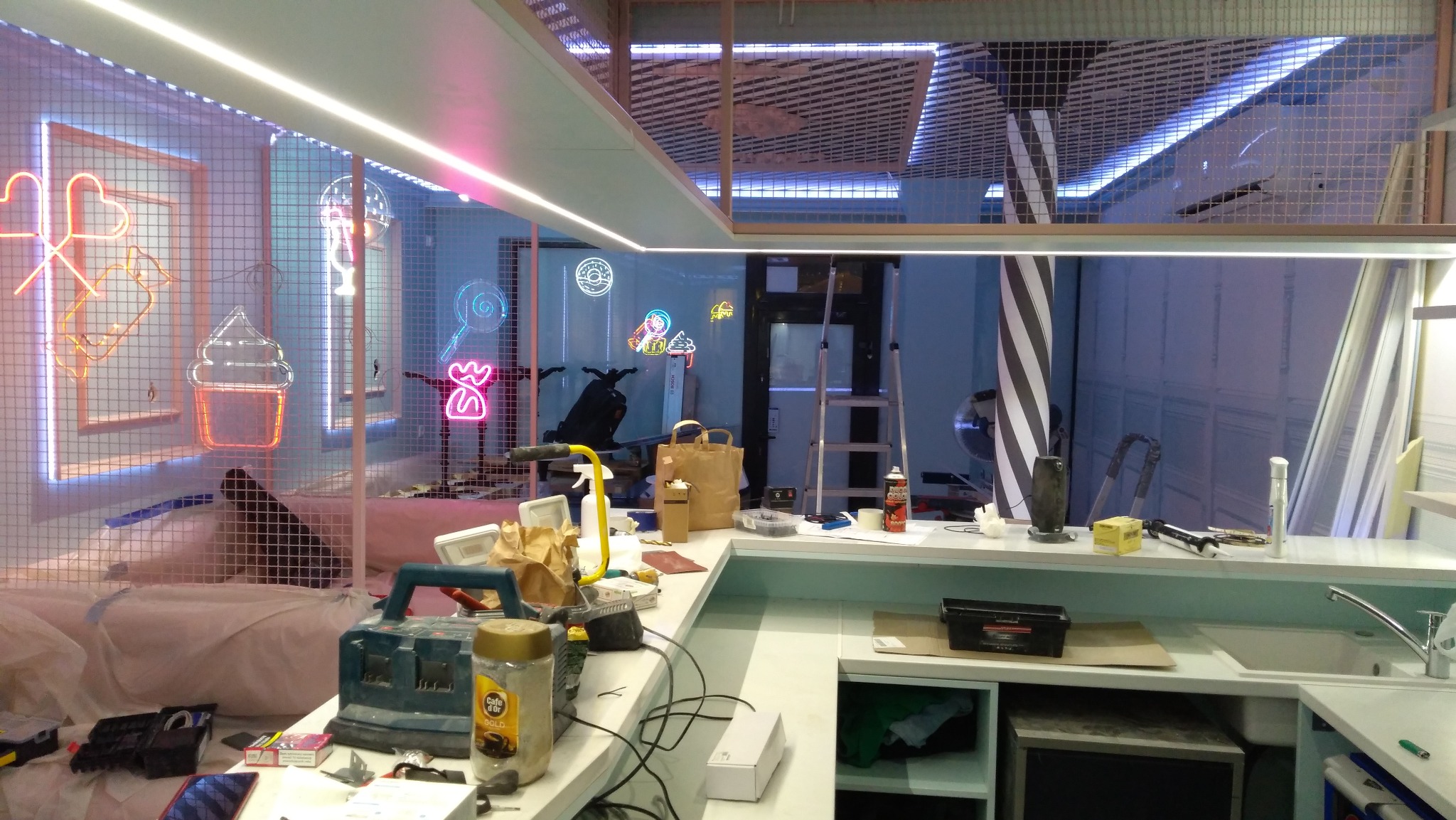 Wnętrze lokalu w trakcie wykańczania z neonami w kształcie słodyczy, narzędziami i materiałami budowlanymi na blacie. Widoczna drabina i oświetlenie LED.