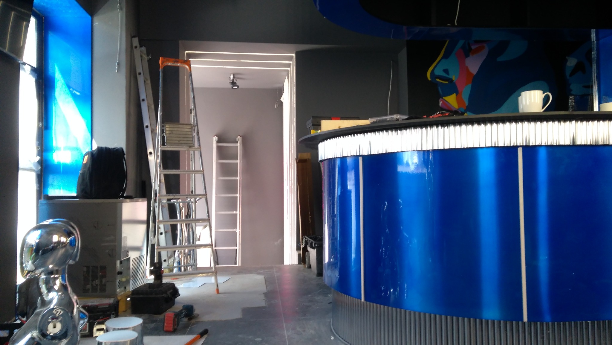 Instalacja oświetlenia LED w lokalu komercyjnym. Widoczne drabiny, narzędzia i fragment baru z niebieskim frontem. Sufit podwieszany z liniami świetlnymi.