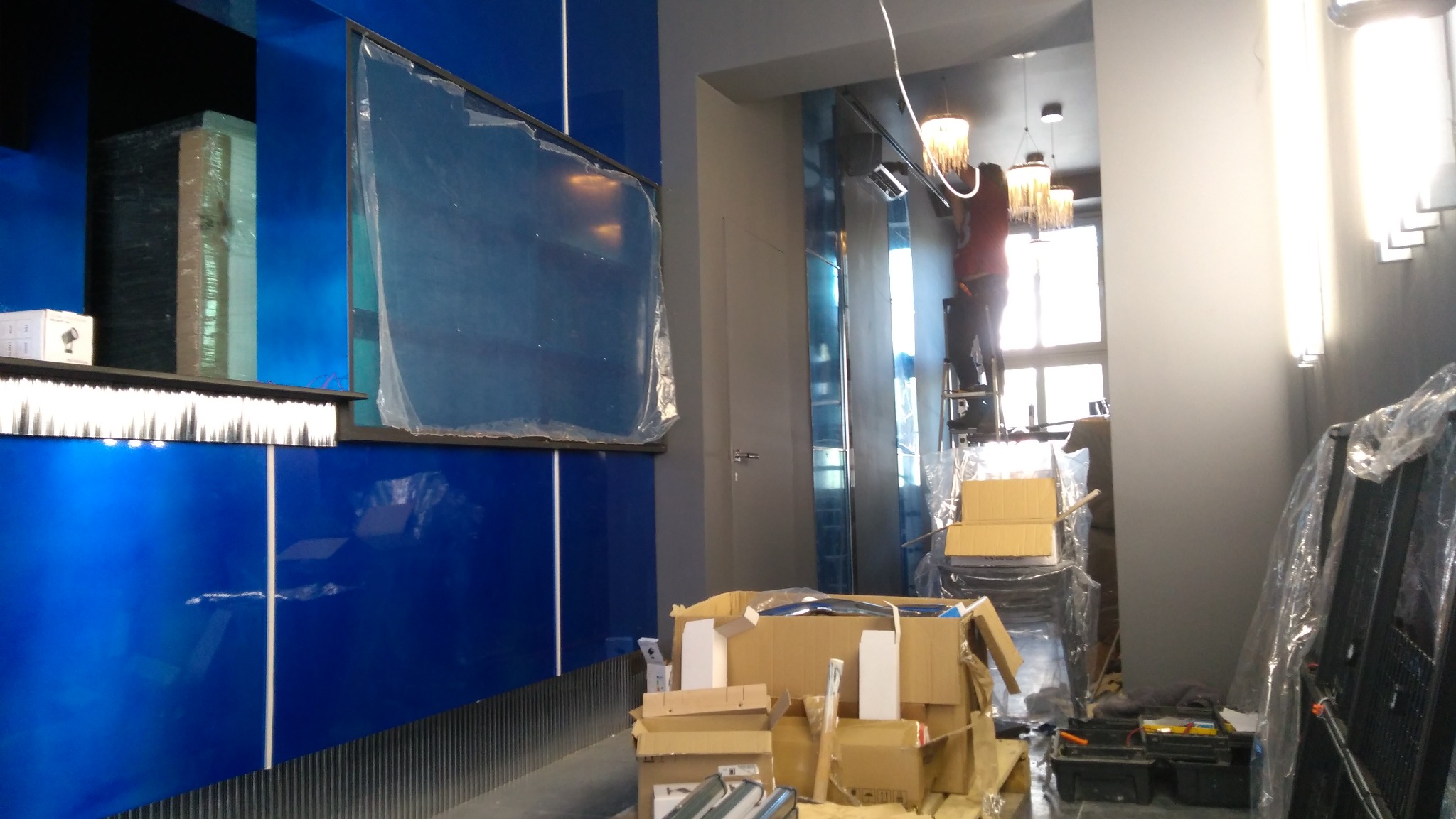 Montaż oświetlenia: instalator na drabinie, w tle lampy wiszące, na pierwszym planie niebieska ściana i kartony z materiałami.