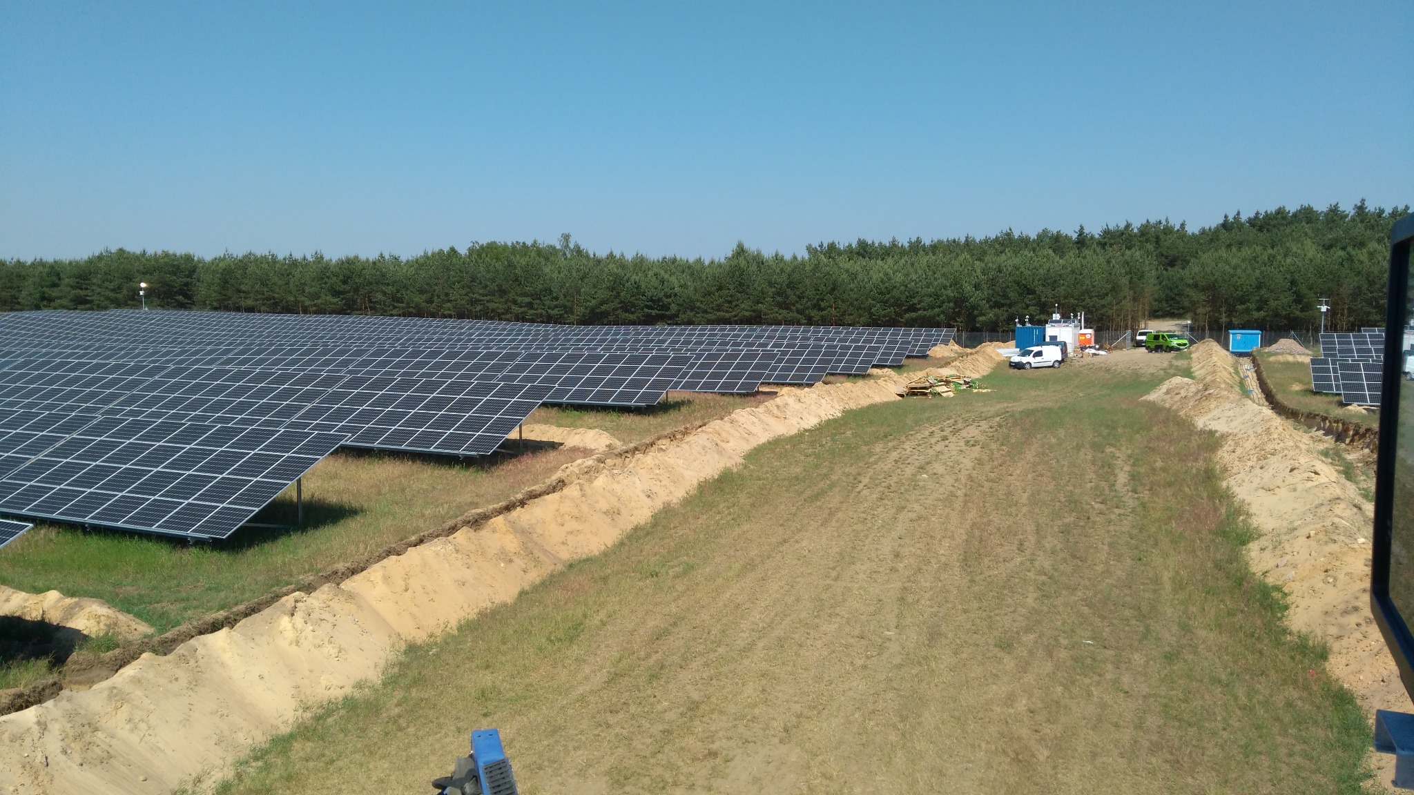 Rozległa farma paneli fotowoltaicznych z wykopami, widok z góry na instalację w słoneczny dzień. W tle las i zabudowania techniczne.