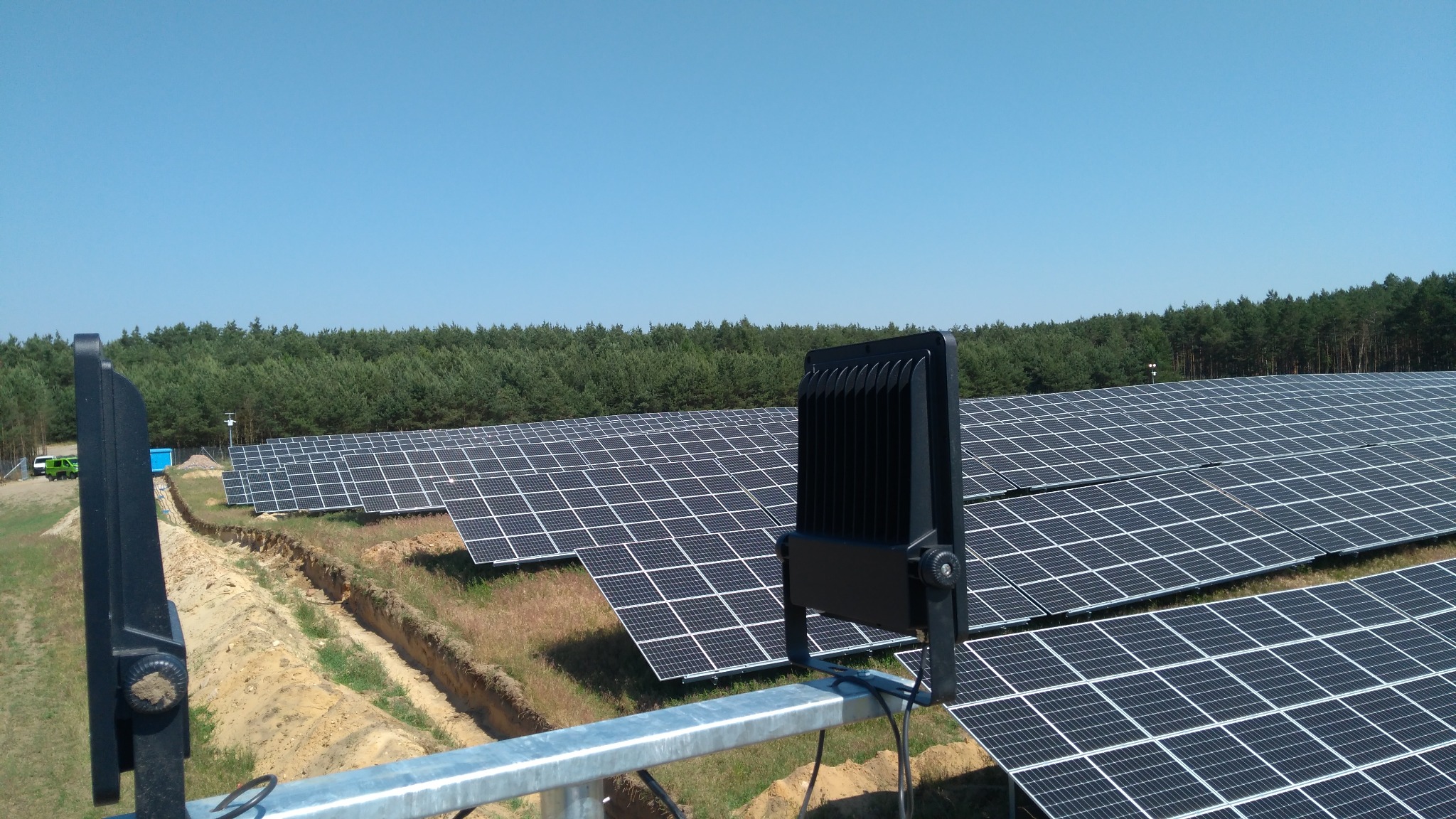 Oświetlenie LED na tle farmy paneli słonecznych i lasu. Kąt ujęcia z góry, widoczne elementy montażowe. Słoneczny dzień, błękitne niebo.