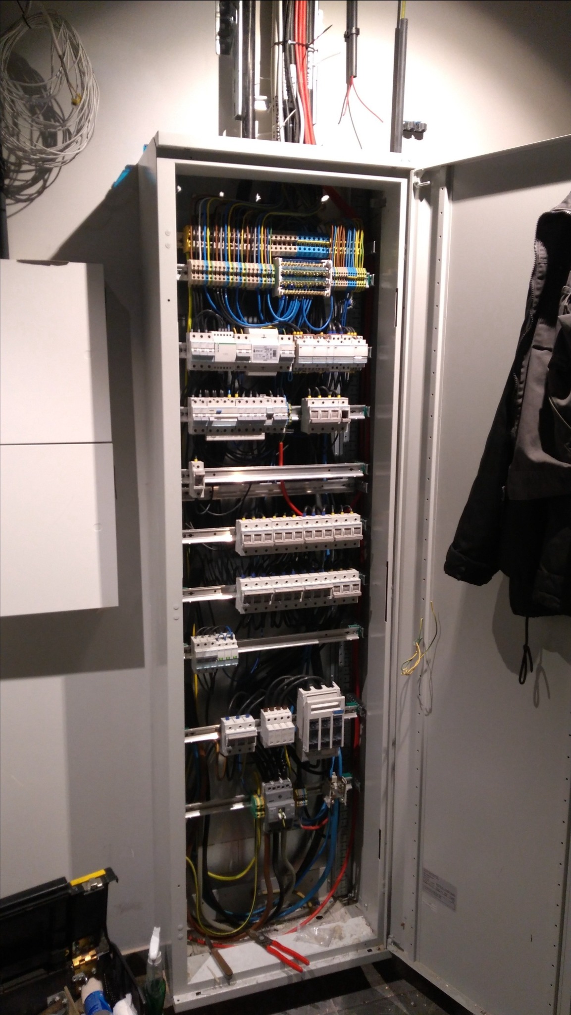 Otwarta skrzynka elektryczna z widocznymi bezpiecznikami, kablami i złączkami. Obok skrzynki narzędzia i czarna kurtka. Wykonana instalacja elektryczna.