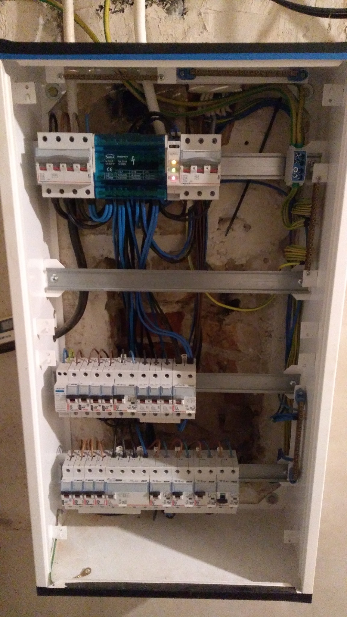 Wnętrze białej skrzynki rozdzielczej z bezpiecznikami i przewodami. Widoczne liczne kable w kolorach niebieskim, żółtym i czarnym oraz metalowe szyny montażowe.