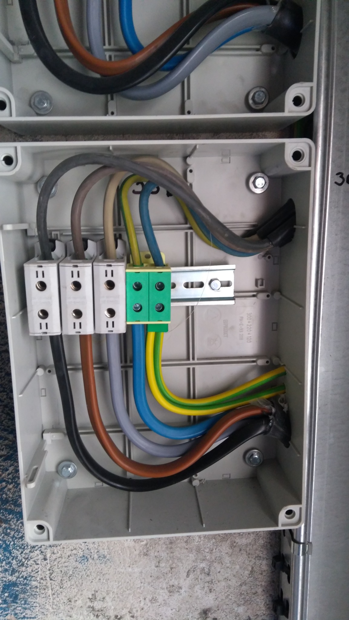 Wnętrze puszki elektrycznej z podłączonymi przewodami o różnych kolorach. Widoczne złącza, listwa montażowa i śruby mocujące. Detal instalacji w szarej obudowie.