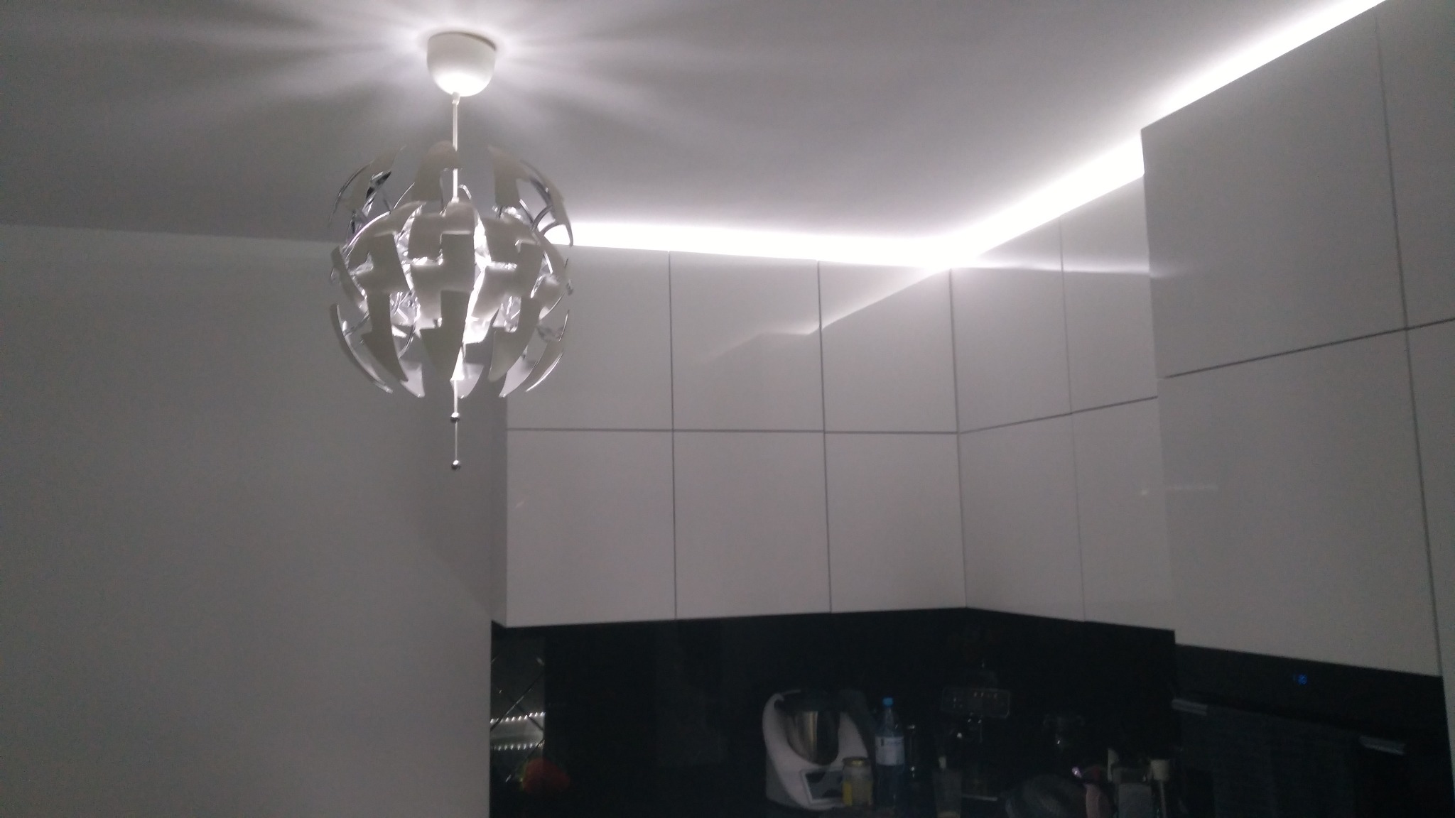 Nowoczesne oświetlenie LED podszafkowe w kuchni z białymi frontami i designerską lampą wiszącą, tworzące jasną i minimalistyczną przestrzeń.