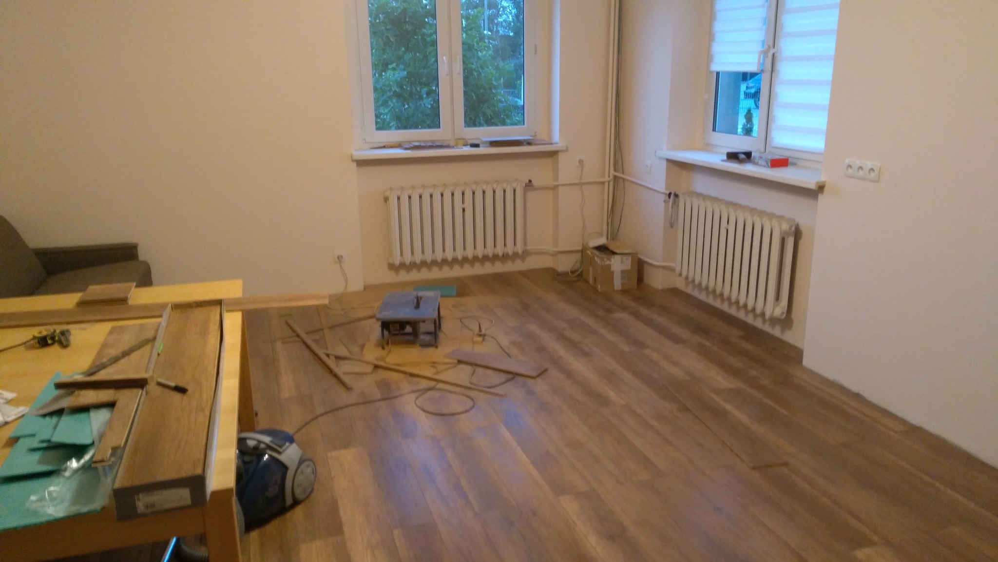 Układanie paneli podłogowych w pokoju z dwoma oknami i grzejnikami. Widoczny stół z narzędziami i odkurzacz. Część podłogi już ułożona.