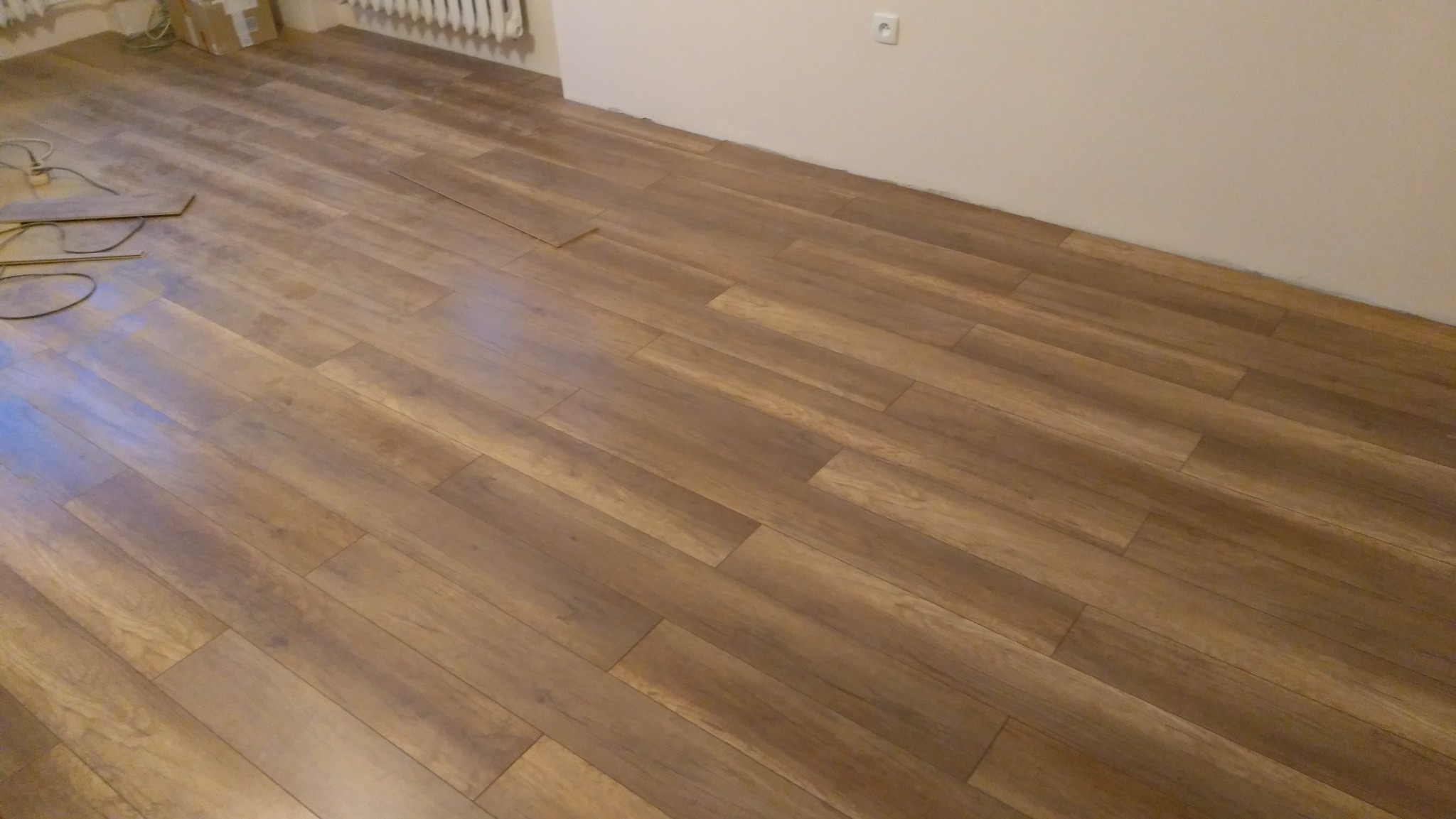 Ułożone panele podłogowe w odcieniach brązu, widoczne kable i fragment radiatora przy ścianie. Jasne ściany i gniazdko elektryczne.