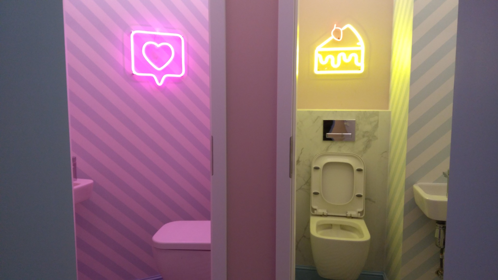 Dwa wejścia do łazienek z neonami: serce na różowo-białych pasach i ciastko na żółto-białych pasach. Widoczne toalety i umywalki.