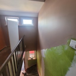 Marek Woch - Klatka schodowa w trakcie remontu: dolna część ściany pomalowana na jaskrawy zielony kolor, górna na beżowo-brązowy, widoczne narzędzie do malowania przy ścianie, drewniana balustrada schodów, okna...