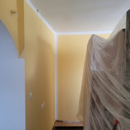 Marek Woch - Korytarz w trakcie malowania na kolor żółty, zabezpieczone meble folią ochronną, widoczne gniazdka elektryczne i białe listwy przypodłogowe.