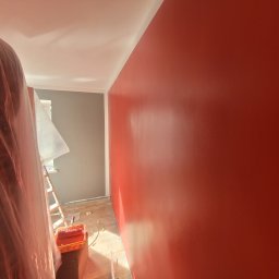 Marek Woch - Świeżo pomalowana na czerwono ściana z widocznymi smugami po wałku, zabezpieczona podłoga i drabina w tle, przygotowania do malowania szarej ściany w pokoju.