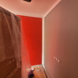 Marek Woch - Świeżo pomalowane ściany w pokoju: jedna czerwona, druga szara, z widocznymi gniazdkami elektrycznymi i zabezpieczonymi meblami folią malarską, na podłodze leżą narzędzia malarskie.