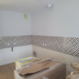 Marek Woch - Fragment kuchni w trakcie prac wykończeniowych: częściowo położone płytki dekoracyjne z geometrycznym wzorem, zabezpieczone folią meble kuchenne, widoczne punkty instalacji elektrycznej i wodnej.