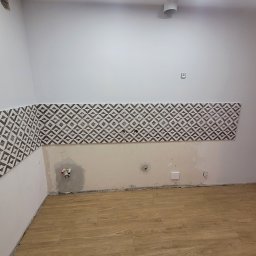 Marek Woch - Fragment ściany w trakcie układania płytek z geometrycznym wzorem, widoczne instalacje hydrauliczne i elektryczne, surowy stan wykończenia, parkiet na podłodze.