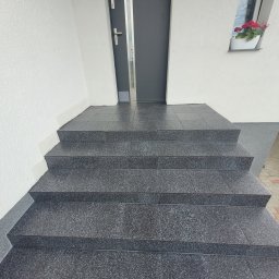 Marek Woch - Zewnętrzne schody wejściowe wyłożone ciemnymi, błyszczącymi płytkami z delikatnym wzorem, prowadzące do szarych drzwi wejściowych z szybą i kwiatów w doniczce na parapecie okna.