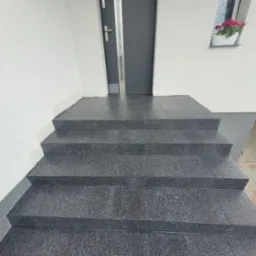 Zewnętrzne schody wejściowe wyłożone ciemnymi, błyszczącymi płytkami z delikatnym wzorem, prowadzące do szarych drzwi wejściowych z szybą i kwiatów w doniczce na parapecie okna.