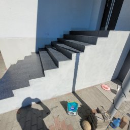 Marek Woch - Wykończone płytkami schody zewnętrzne prowadzące do domu, z widocznym workiem kleju Atlas i narzędziami na brukowanym podłożu, cień osoby w kadrze.