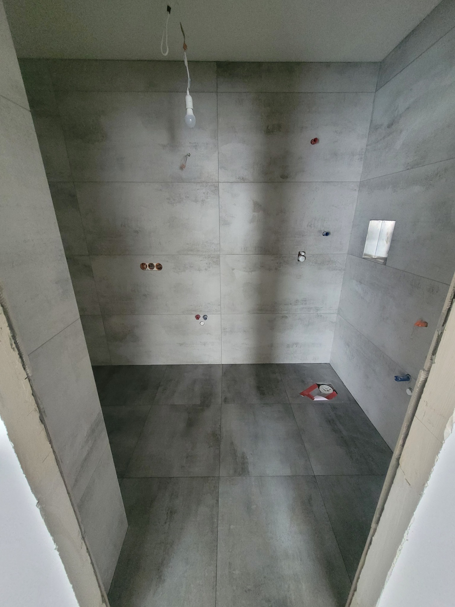 Wnętrze łazienki w trakcie remontu, widoczne szare płytki imitujące beton na ścianach i podłodze, wystające instalacje wodno-kanalizacyjne, surowa żarówka zwisająca z sufitu i osłona odpływu...