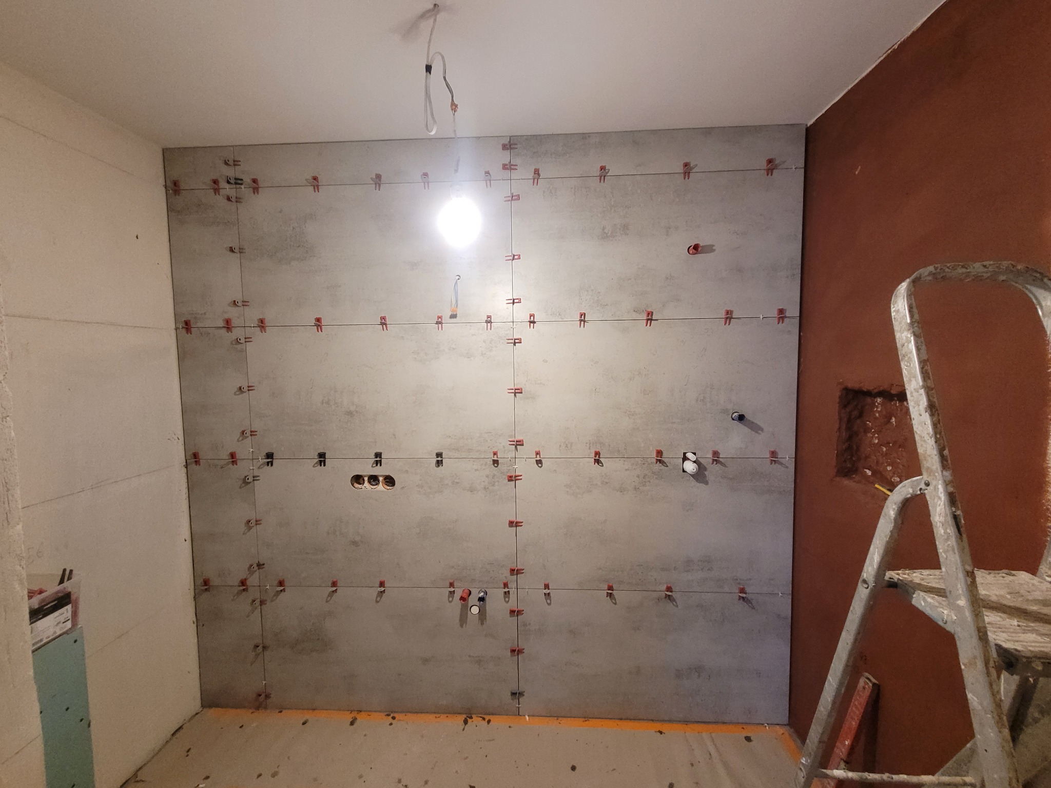 Ściana w trakcie układania płytek imitujących beton, z użyciem systemu poziomowania, widoczne otwory na gniazdka elektryczne i drabina malarska w tle.