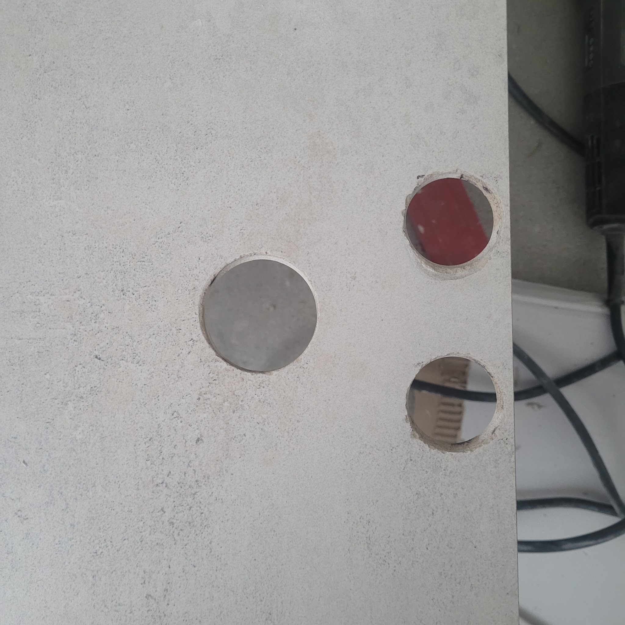 Płyta gipsowa z trzema wywierconymi otworami, jeden z widoczną czerwoną ścianą w tle, obok widoczny fragment wiertarki z kablem.
